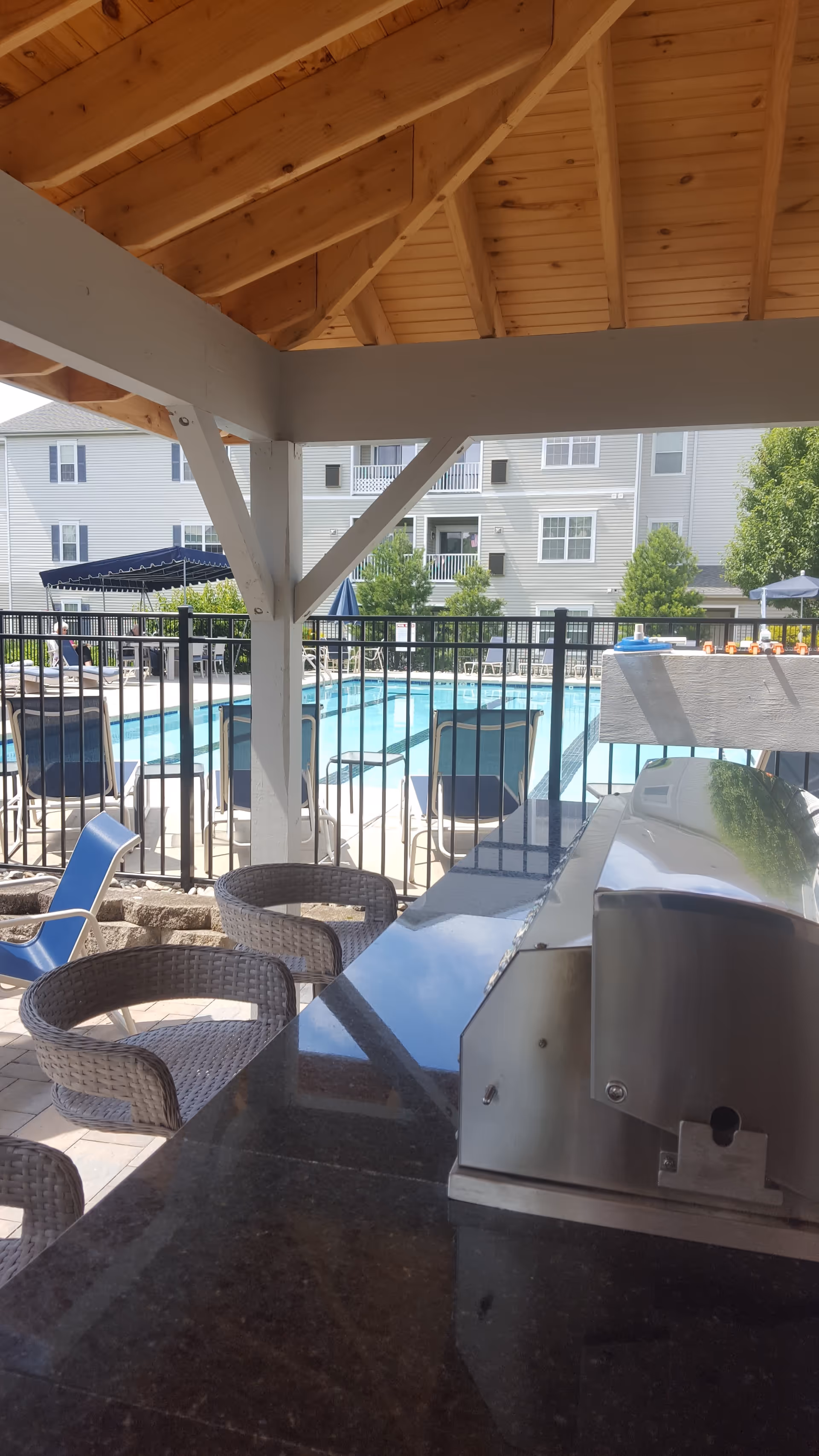 Outdoor covered seating area with wicker chairs and a granite countertop next to a stainless steel grill, overlooking a fenced swimming pool with lounge chairs and umbrellas, with a multi-story residential building in the background.