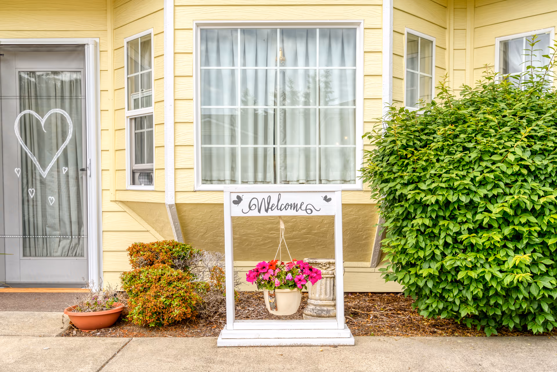 Exterior view of a yellow building with a large window and a glass door decorated with a white heart and small hearts. In front of the window, there is a white wooden stand with a hanging pot of pink flowers and a sign that reads 'Welcome'. There are green bushes and a small potted plant near the entrance.
