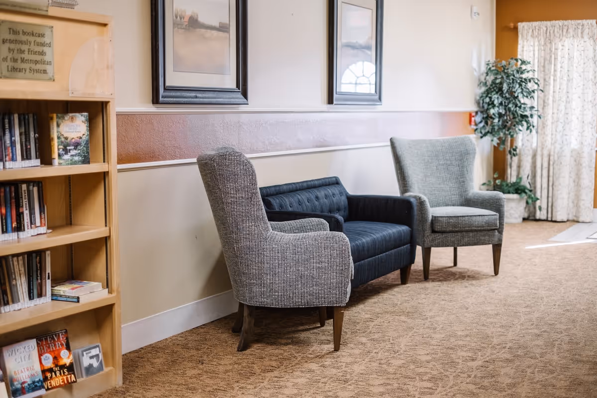 A cozy seating area in a senior living facility with two gray upholstered armchairs and a dark blue tufted loveseat arranged against a beige and brown wall. To the left, there is a wooden bookcase filled with books, and on the right side, a potted plant is placed near a window with patterned curtains allowing natural light to enter.
