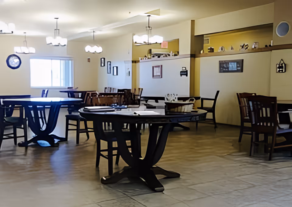 Interior view of a dining room in an assisted living facility with several round and rectangular wooden tables and chairs arranged neatly. The room is well-lit with multiple ceiling light fixtures and has a window on one side. The walls are decorated with framed pictures and small shelves holding decorative items.