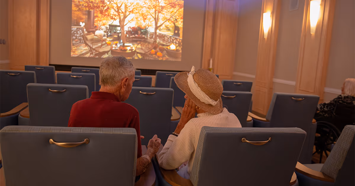 Two elderly people sitting in a small theater or screening room with blue cushioned chairs, watching a large screen displaying an autumn outdoor scene with trees and pumpkins.