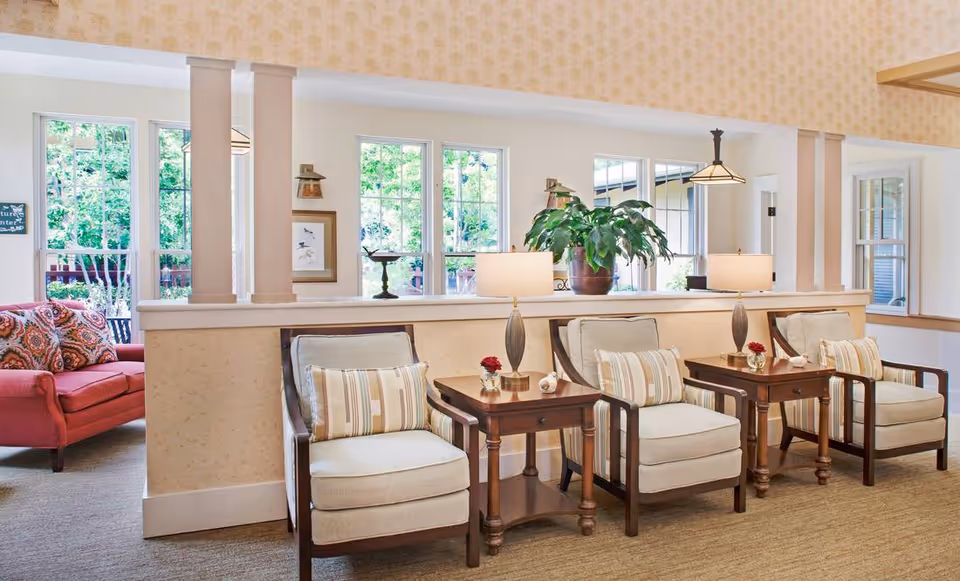 Bright communal lounge area with upholstered armchairs, wooden side tables and lamps facing large windows and a red sofa in the background.