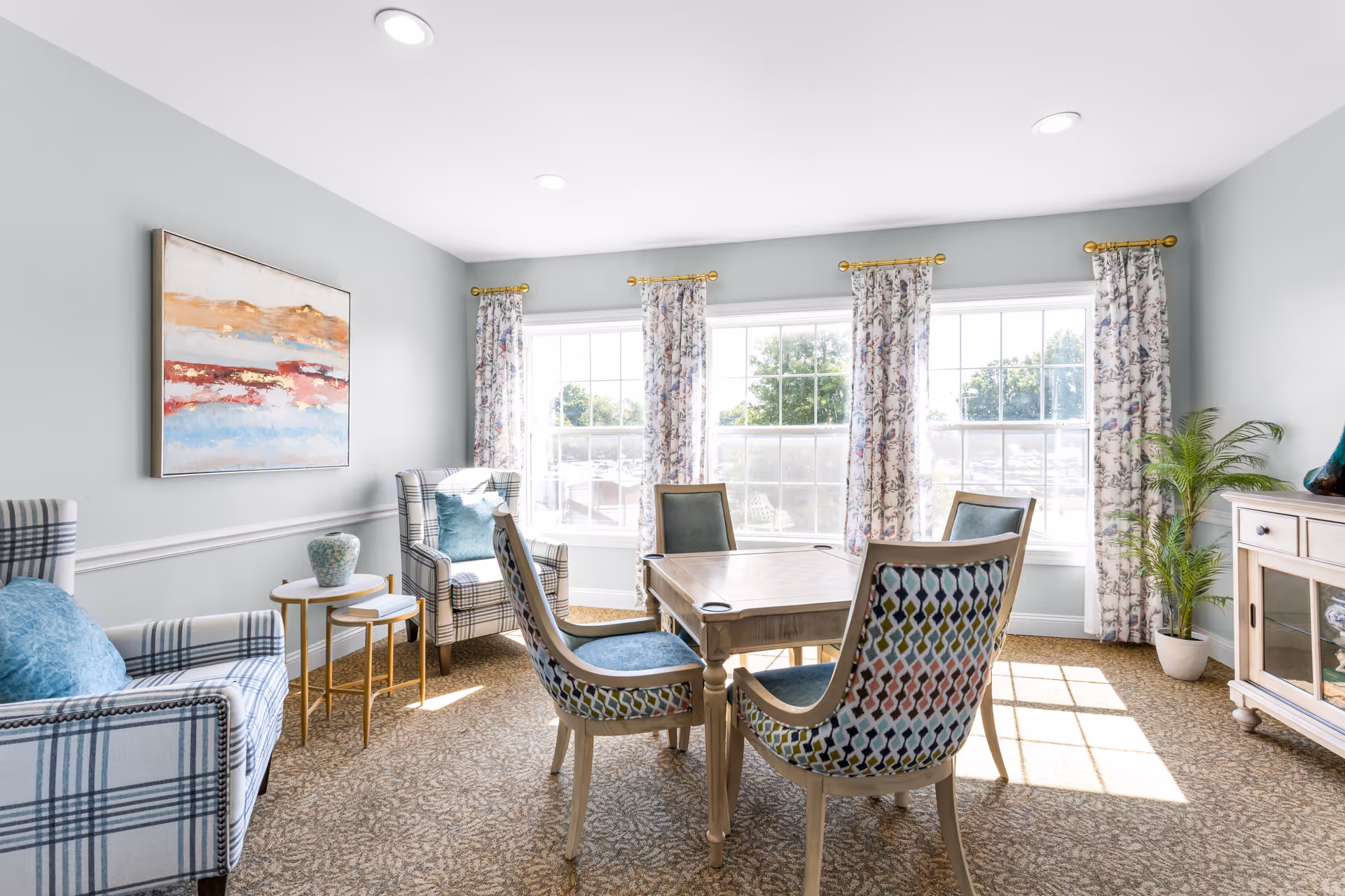 A bright and cozy sitting area with a square wooden table surrounded by four upholstered chairs with patterned backs and blue seats. Two plaid armchairs with blue cushions are positioned near a small round side table with a decorative vase. Large windows with floral curtains allow natural light to fill the room, and a colorful abstract painting hangs on the light gray wall. A potted plant and a wooden cabinet with glass doors are also visible.