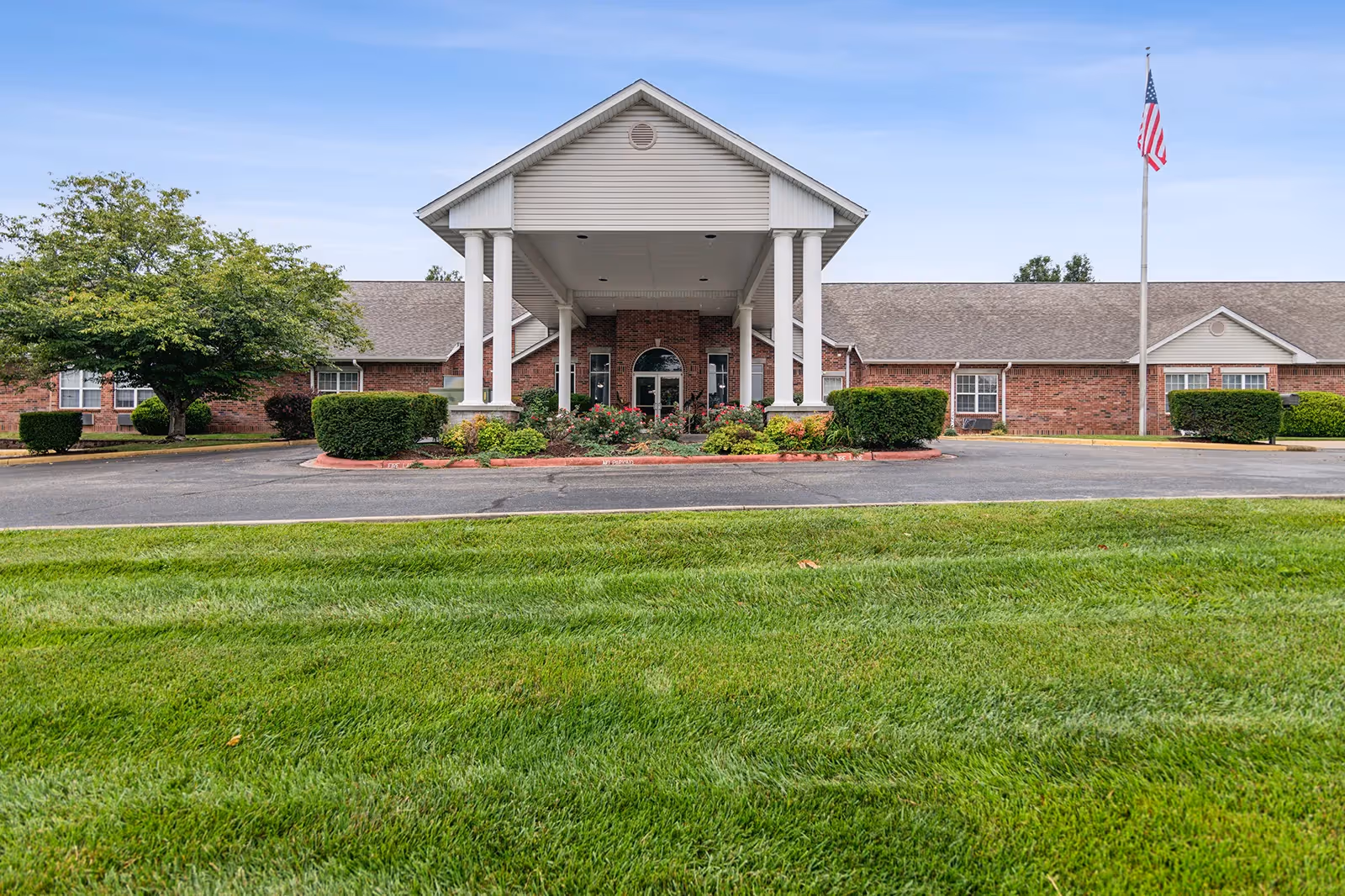 Front entrance of a single-story brick senior living building with a covered portico, landscaped shrubs, and an American flag.