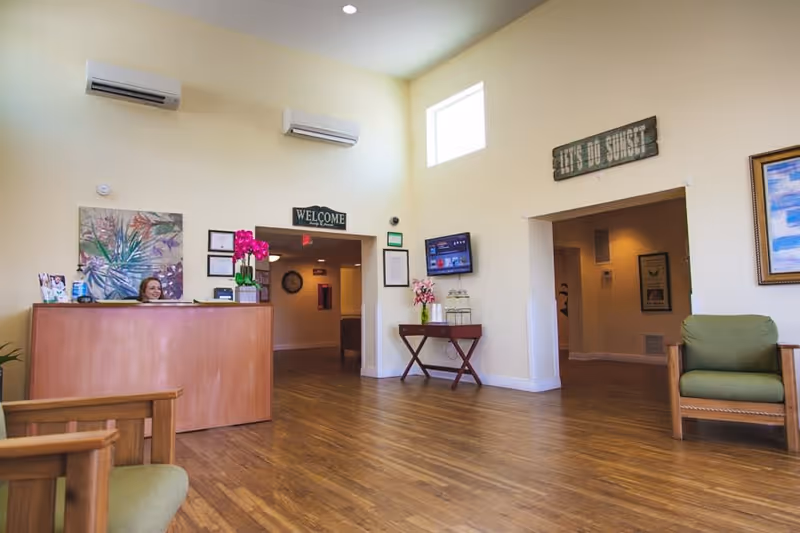 Reception area of a senior living facility with a wooden desk where a woman is seated, wooden chairs with green cushions, light yellow walls, a wooden floor, wall-mounted air conditioning units, a welcome sign above a doorway, a small table with flowers and water dispensers, and framed artwork on the walls.
