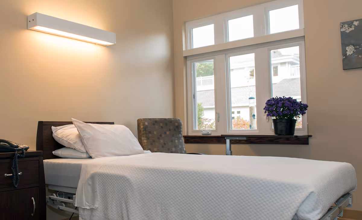 A neatly made single bed with white bedding and pillows in a well-lit room. Next to the bed is a dark wooden nightstand with a telephone on it. A patterned armchair is positioned near a window with multiple panes, through which an exterior building and greenery are visible. A potted plant with purple flowers sits on the windowsill. The walls are painted a light beige color, and a rectangular light fixture is mounted above the bed.