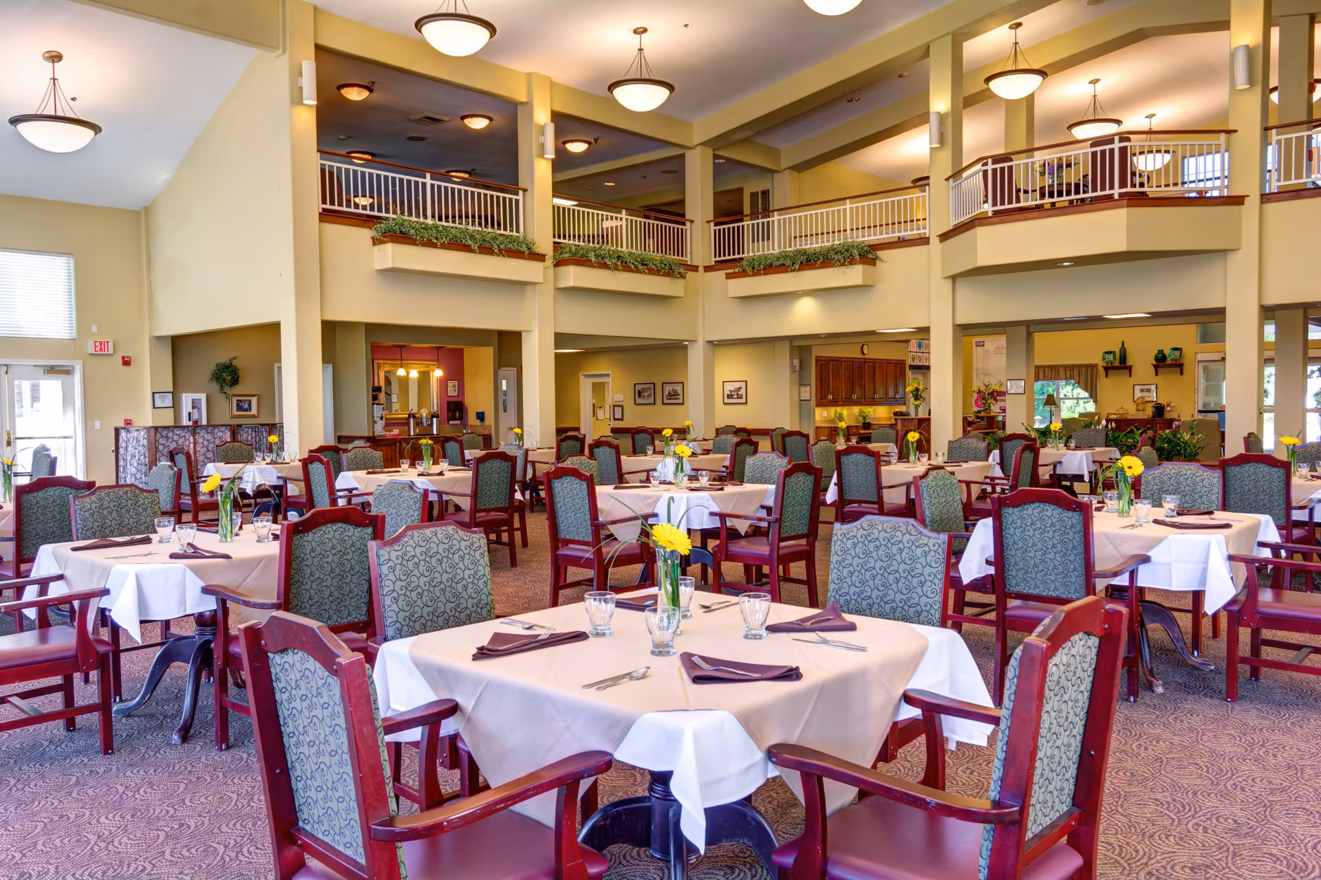 A spacious dining room with multiple tables covered in white and beige tablecloths, each set with glasses, napkins, and yellow flowers in vases. The room features high ceilings with hanging light fixtures, a second-floor balcony with railings, and a warm, inviting atmosphere.