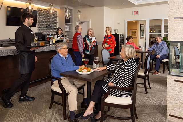 A group of elderly people socializing and dining in a well-lit common area with tables and chairs. A waiter is serving drinks at a bar counter, and a bartender is behind the counter. The room has modern lighting fixtures, a TV mounted on the wall, and a fireplace on the right side.