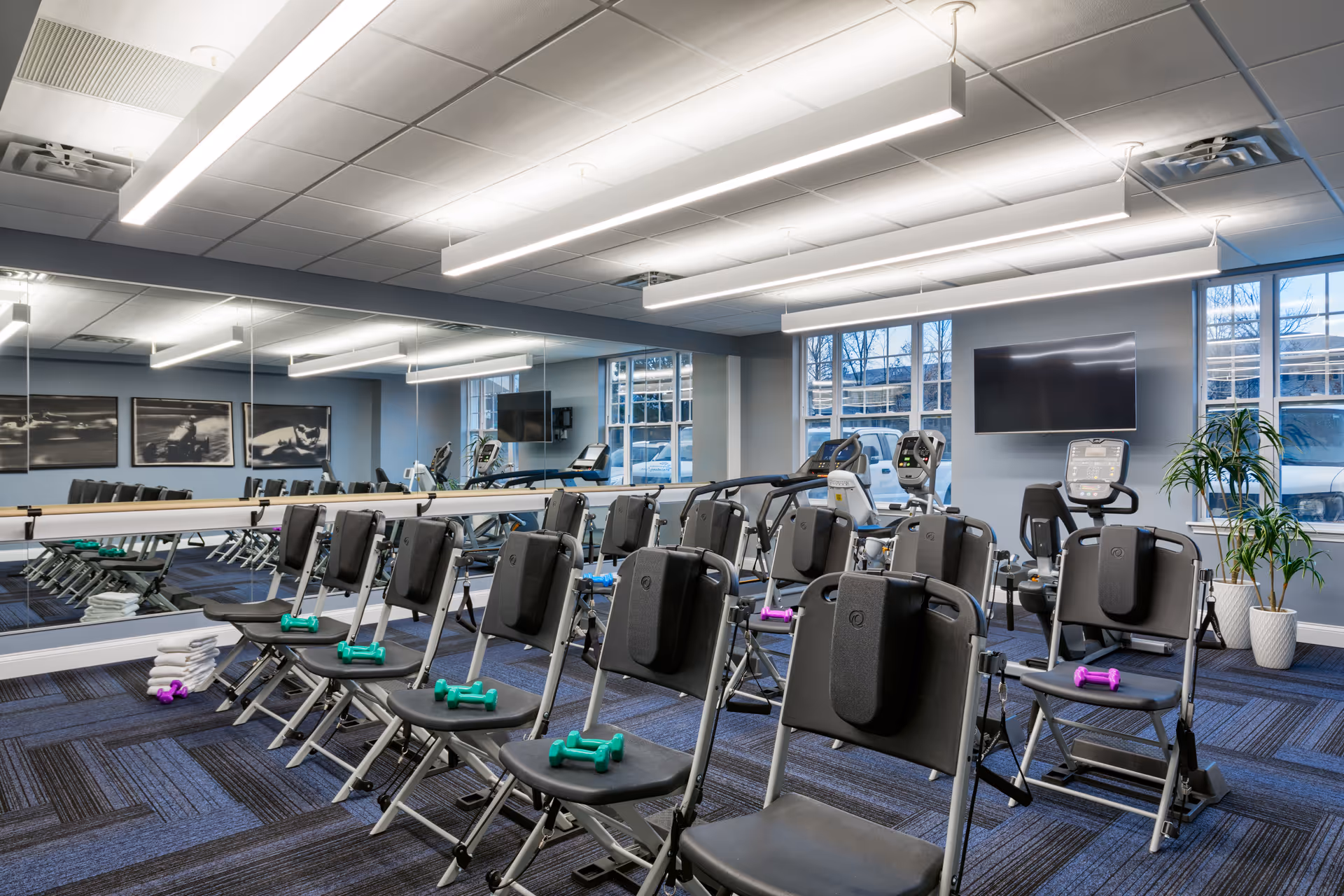 A fitness room with multiple black chairs arranged in rows, each chair holding small colorful dumbbells. The room has large mirrors on one wall, exercise equipment including stationary bikes and treadmills near the windows, and a flat-screen TV mounted on the wall. The ceiling has modern linear lights, and there are potted plants near the windows.