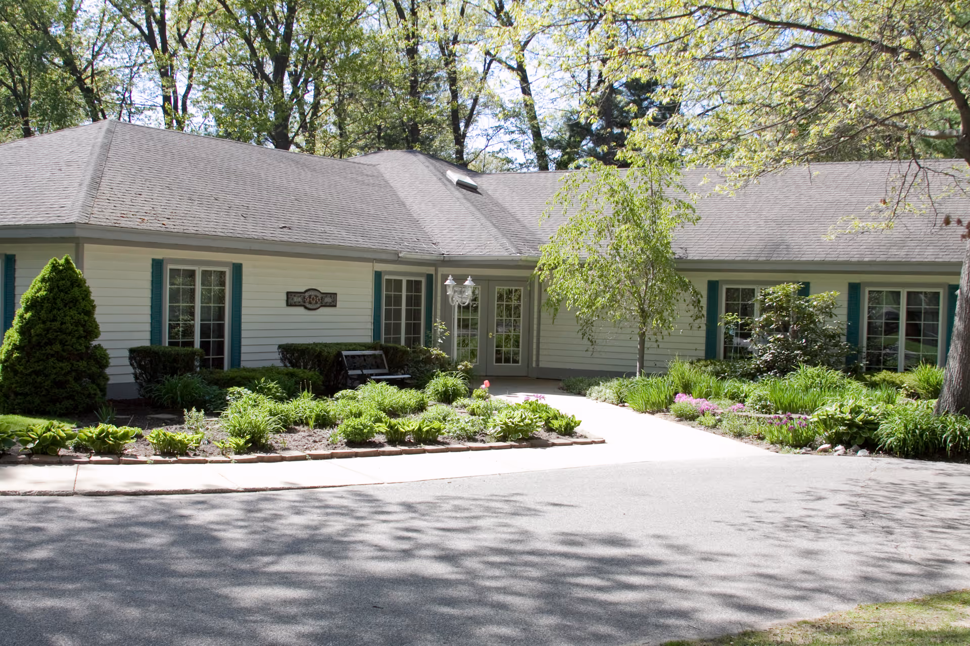 Exterior view of a single-story building with white siding and teal window shutters, surrounded by well-maintained landscaping including bushes, flowers, and trees. A paved driveway and sidewalk lead to a set of double glass doors at the entrance.