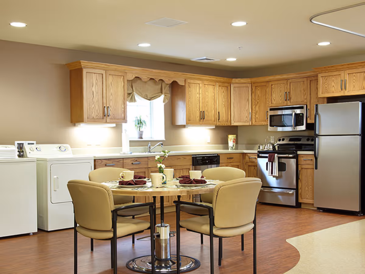 A bright kitchen area with wooden cabinets, stainless steel refrigerator, stove, and microwave. There is a round dining table with four beige cushioned chairs, set with plates, cups, and napkins. A washer and dryer are visible on the left side under a window with a small plant on the windowsill.