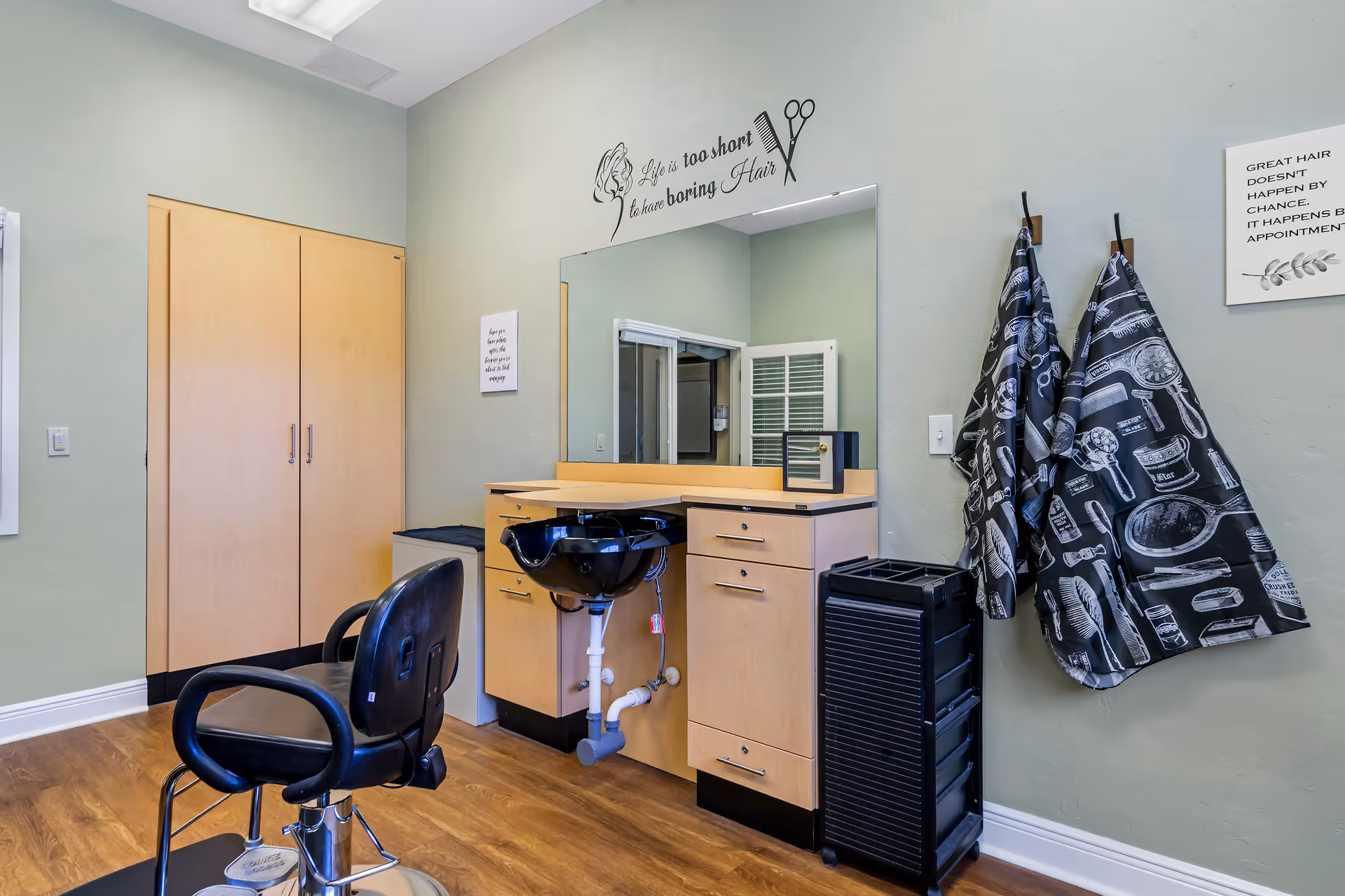 Small hair salon station with a styling chair, wash basin, mirror, cabinets and hanging capes in a room.