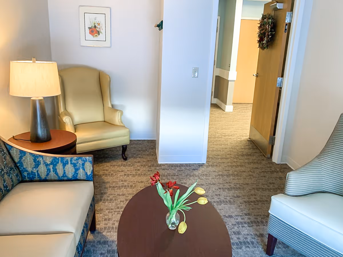 Small sitting room with armchairs, a sofa, a coffee table with flowers, a lamp, and an open door to a hallway.