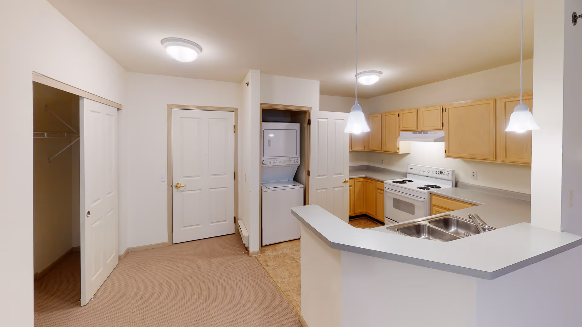 Interior view of a senior community apartment kitchen area with light wood cabinets, a white stove, a double sink on a countertop peninsula, and two pendant lights. Adjacent to the kitchen is a laundry closet with a stacked washer and dryer. There is a carpeted area with a closet on the left and a closed door in the center.