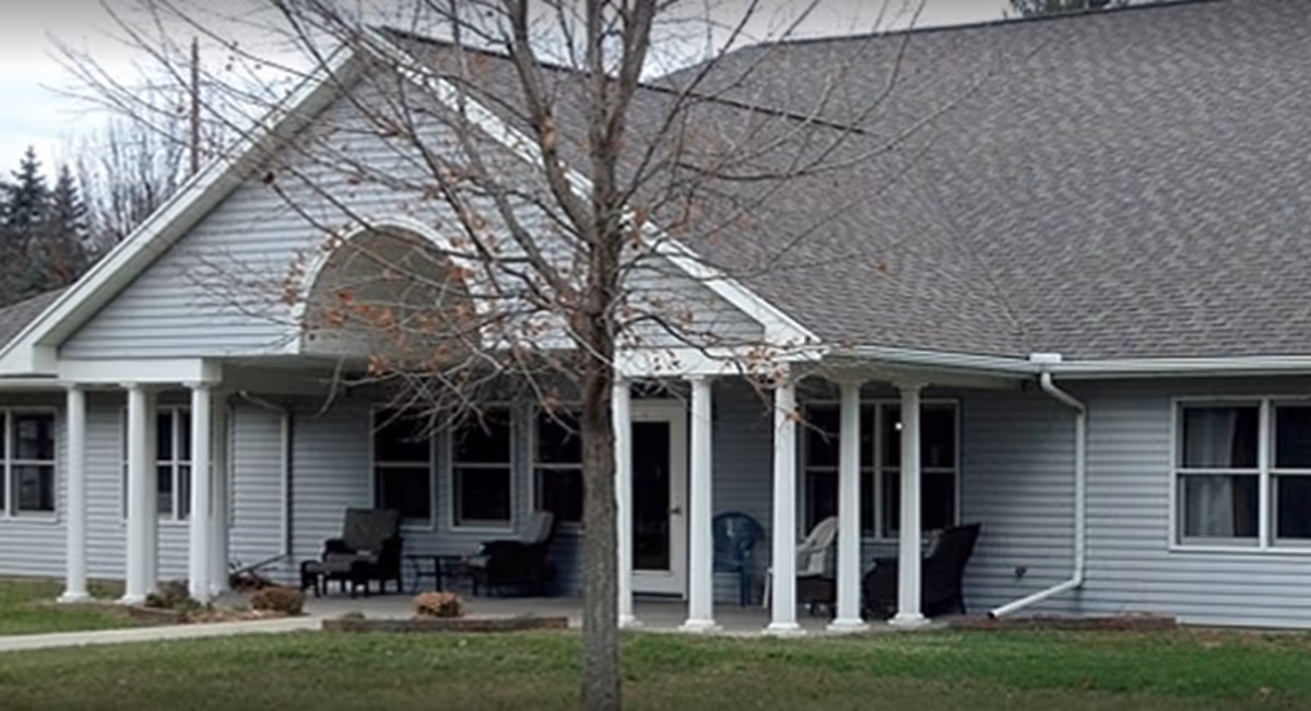 Front exterior of a single-story senior living facility with a covered porch supported by white columns, outdoor chairs, and a leafless tree in front.