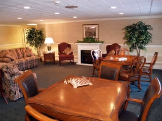 Cozy communal living room with wooden game tables, upholstered chairs and sofas, potted plants, and a fireplace.