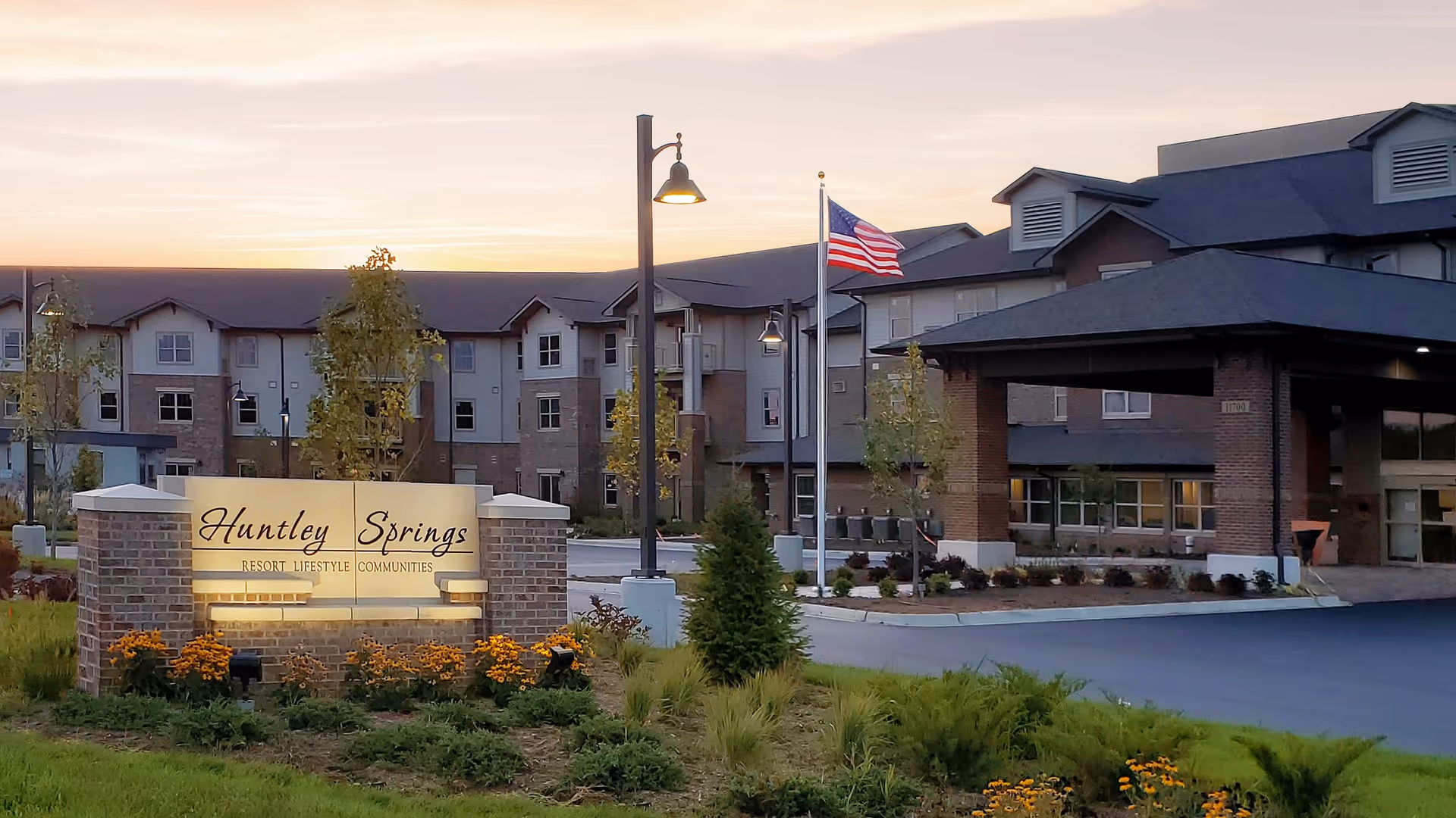 Exterior view of Huntley Springs Retirement Resort at dusk, showing the main entrance with a covered drop-off area, an American flag on a flagpole, landscaped greenery, and a lit sign displaying the facility's name.