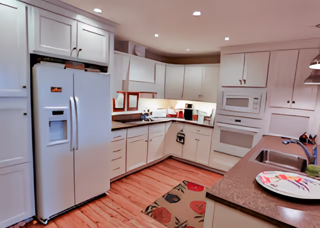 A spacious kitchen with white cabinets and drawers, a double-door refrigerator with an ice dispenser, a built-in microwave and oven, a double sink, and a countertop with a decorative plate. The floor is wooden with a colorful rug near the sink area. The kitchen is well-lit with recessed ceiling lights.
