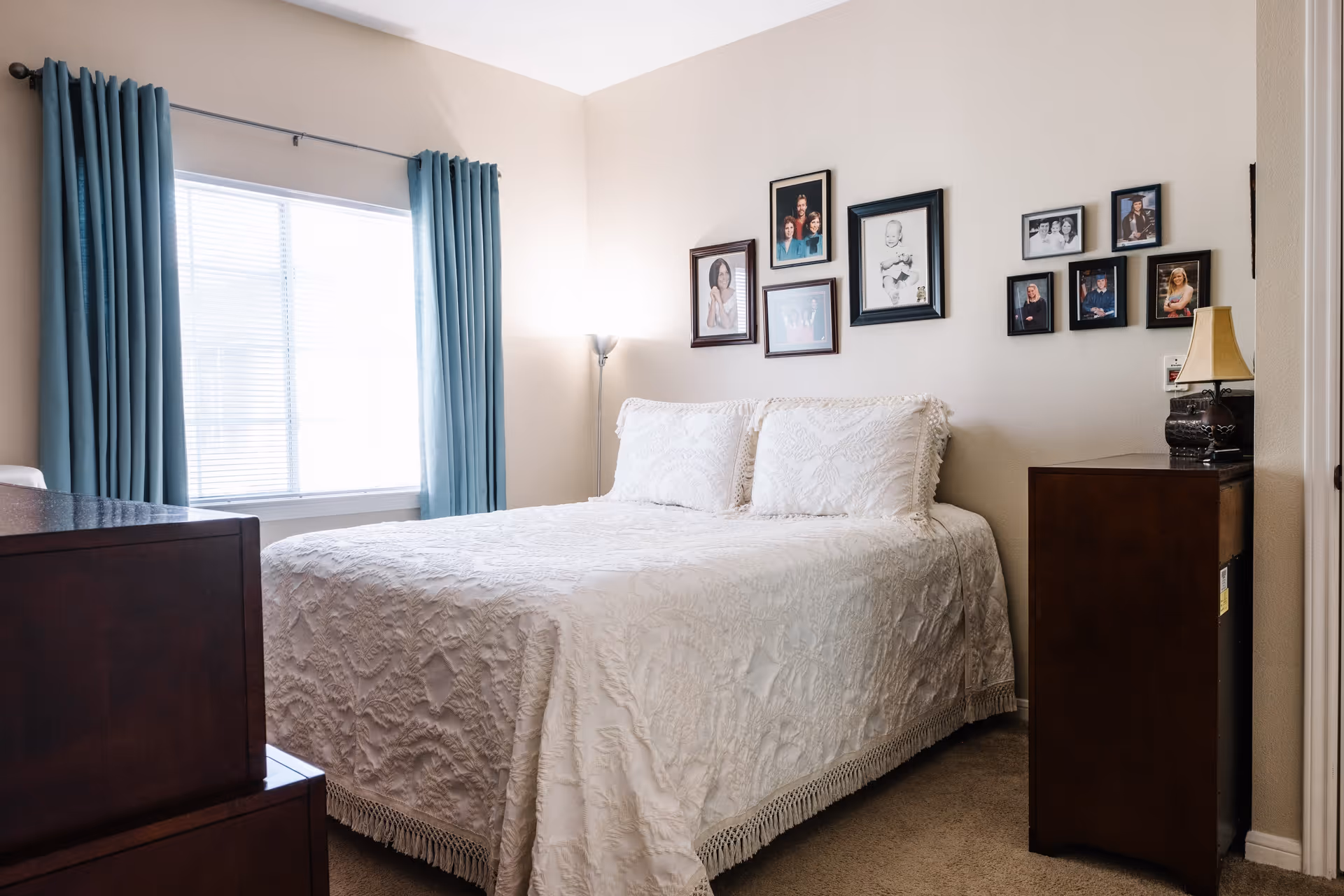 A cozy bedroom with a neatly made bed covered in a white textured bedspread. The bed is positioned against a wall decorated with multiple framed family photos. To the right of the bed is a dark wooden dresser with a small lamp on top. A window with blue curtains allows natural light to brighten the room.