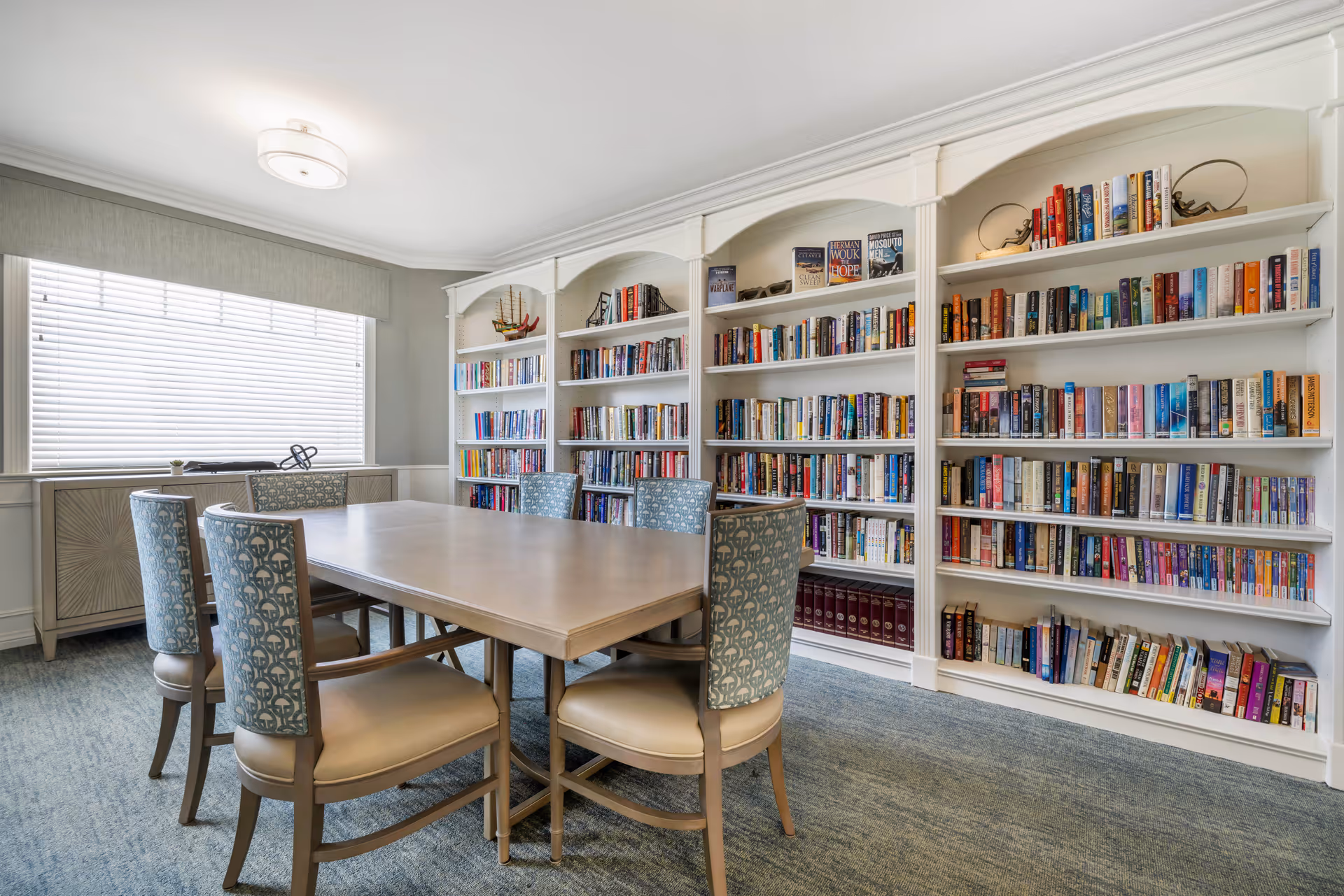 A bright room with a large window covered by blinds, featuring a rectangular wooden table surrounded by six upholstered chairs. Along one wall, there are built-in white bookshelves filled with a variety of books and decorative items. The room has a carpeted floor and a ceiling light fixture.