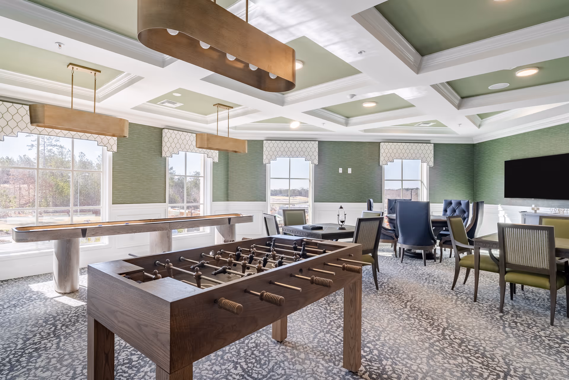 A bright common room with a foosball table, shuffleboard, dining tables and lounge chairs under coffered ceilings.