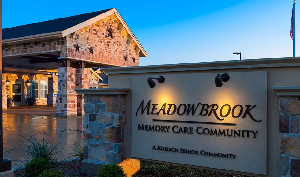 Entrance of Meadowbrook Memory Care Community featuring a stone and brick building with a covered driveway and a large sign illuminated by two lights, displaying the facility's name and noting it as a Koelsch Senior Community.