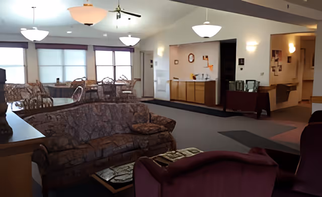Interior view of a senior living facility lounge area with patterned sofas, armchairs, a coffee table, and a dining area with chairs and tables near large windows. The room is softly lit with ceiling lights and wall sconces, and there is a counter area along one wall.