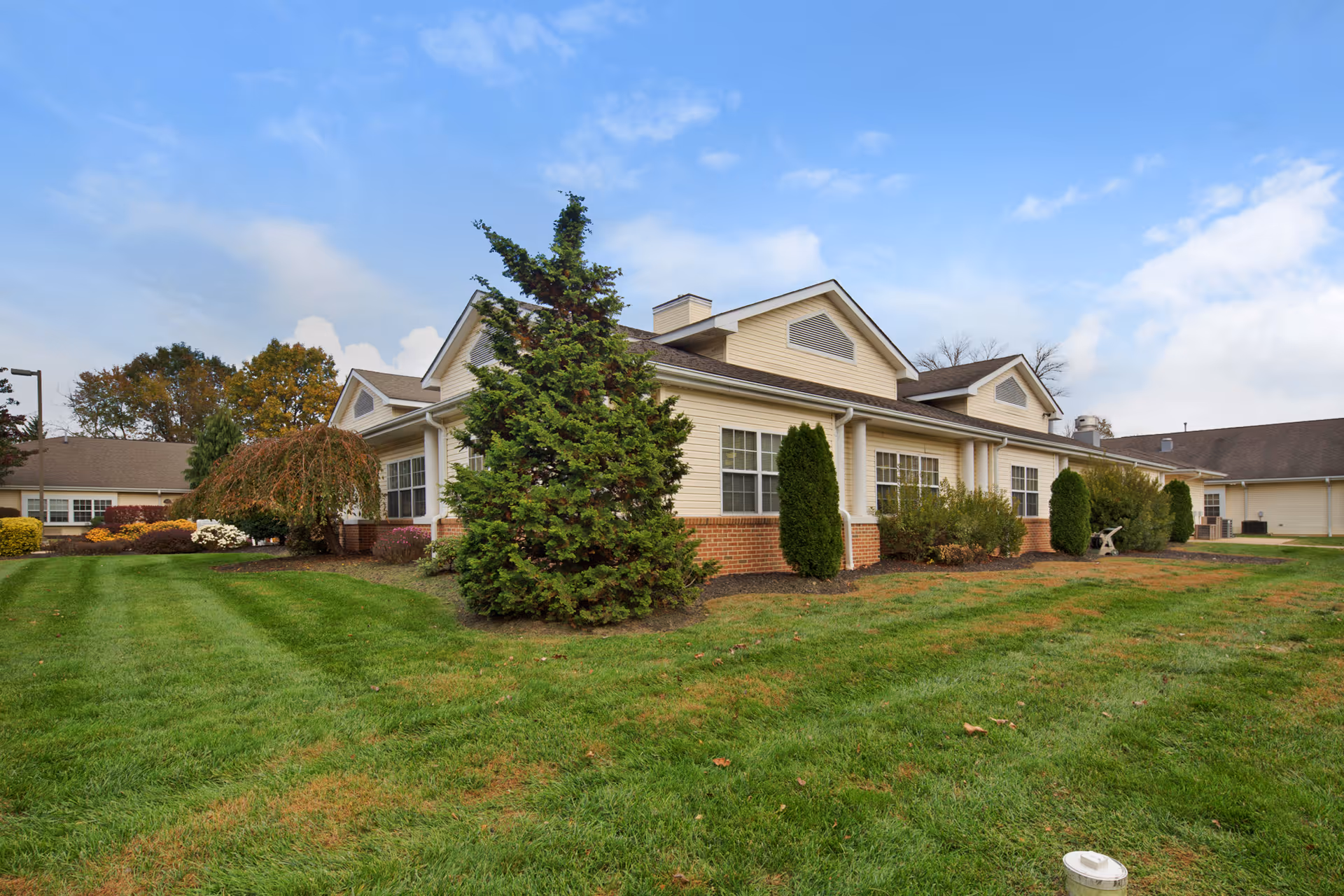 Exterior view of a single-story senior living facility building with beige siding and brick accents, surrounded by green grass, shrubs, and trees under a partly cloudy sky.