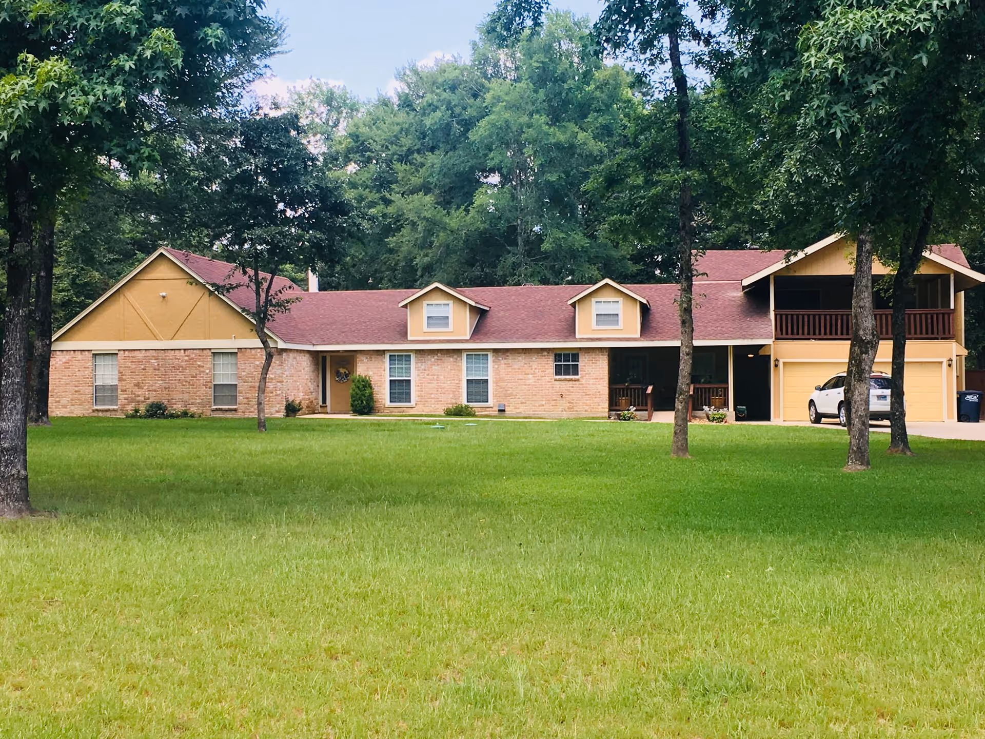 A single-story brick house with a red roof and yellow trim, surrounded by green grass and tall trees. There is a driveway with a white car parked near a garage on the right side of the house.
