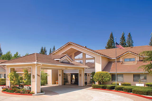 Exterior view of a senior living facility building with a covered entrance, beige walls, and a tiled roof under a clear blue sky. There are well-maintained shrubs, trees, and an American flag on a flagpole near the entrance.