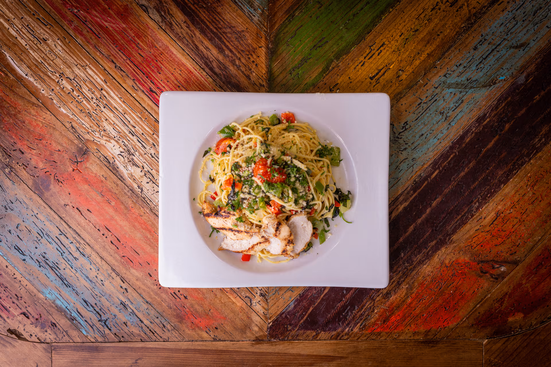 A square white plate of pasta with grilled chicken and vegetables on a colorful wooden table.