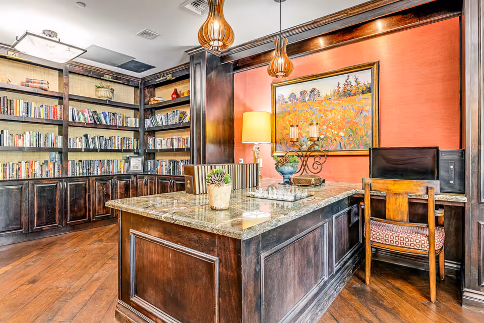 A cozy library room with dark wood bookshelves filled with books, a large wooden table with a marble top in the center, decorative items including a chess set, potted plants, and candles. There is a wooden chair with a patterned cushion in front of a computer on the right side. The walls are painted a warm orange color with a framed painting of a field of flowers hanging above the table. Two pendant lights hang from the ceiling.