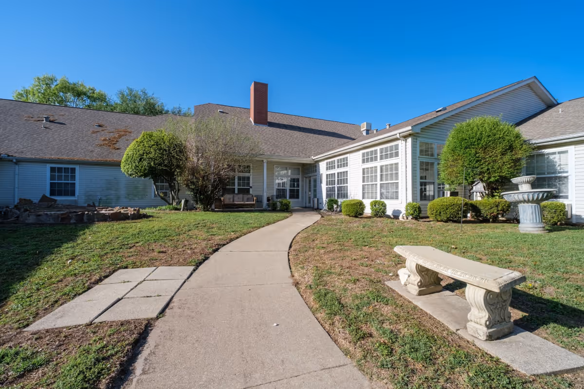 Outdoor view of a senior living facility with a paved walkway leading to the entrance. The building has white siding, multiple windows, and a brown shingled roof. There are trimmed bushes, a stone bench, and a decorative fountain in the grassy area surrounding the walkway under a clear blue sky.