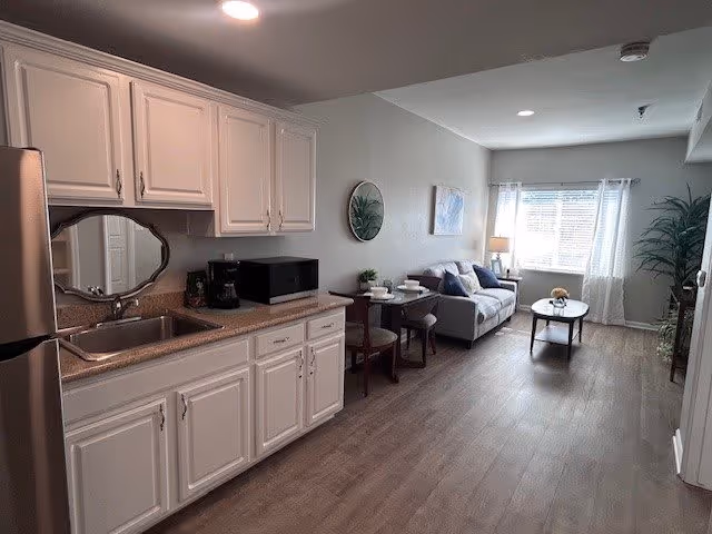 A bright and clean studio apartment interior with a kitchenette on the left featuring white cabinets, a stainless steel sink, a microwave, and a coffee maker. The room extends into a living area with a small round dining table with two chairs, a gray sofa with cushions, a coffee table with a small plant, and a large window with white curtains letting in natural light. There is a potted plant in the corner near the window.