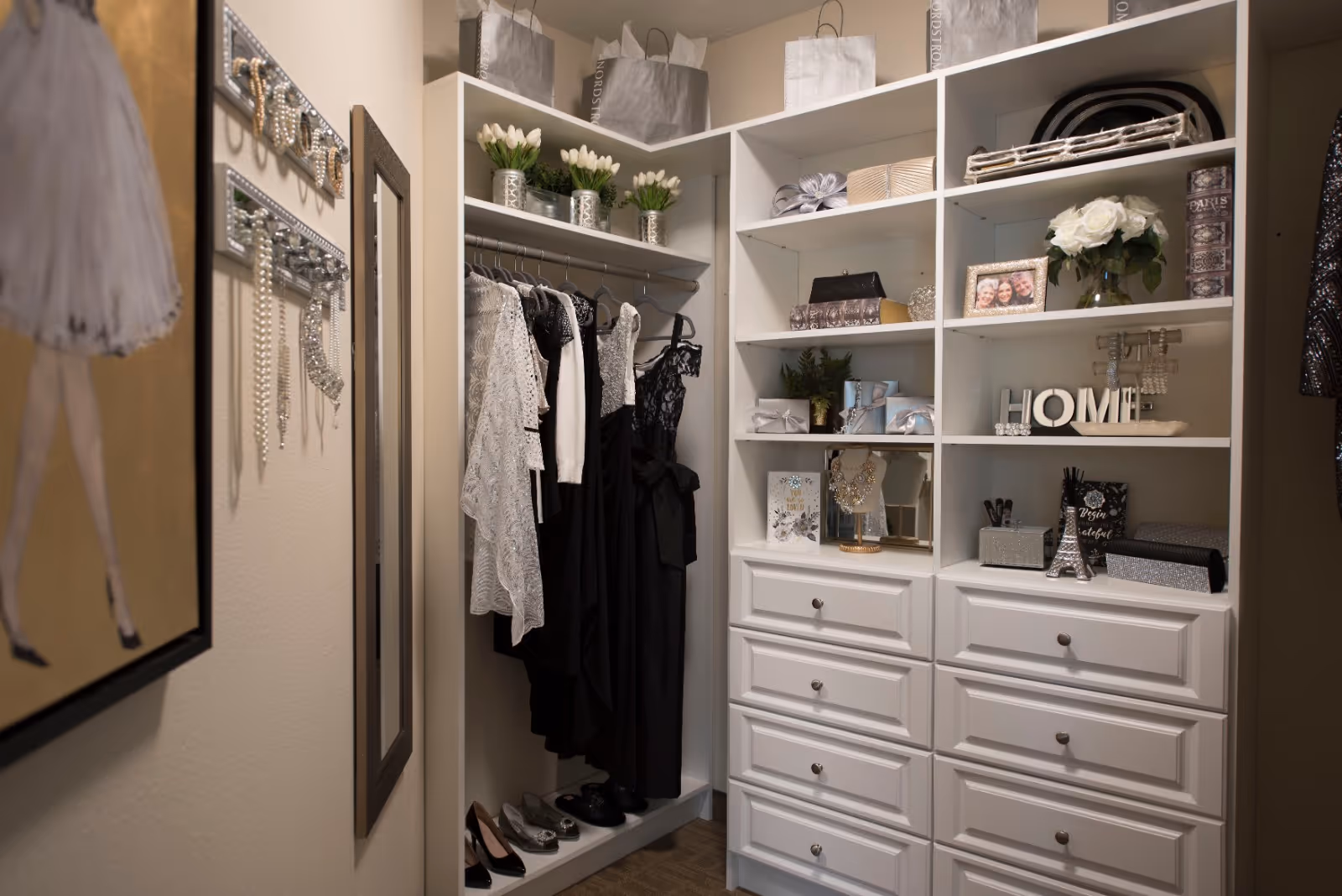 A neatly organized walk-in closet with white shelving and drawers. Several black and white dresses hang on a rod, with shoes placed on the floor below. The shelves hold decorative items including potted plants, gift bags, framed photos, and various accessories. A mirror and a framed ballerina artwork are visible on the adjacent wall.