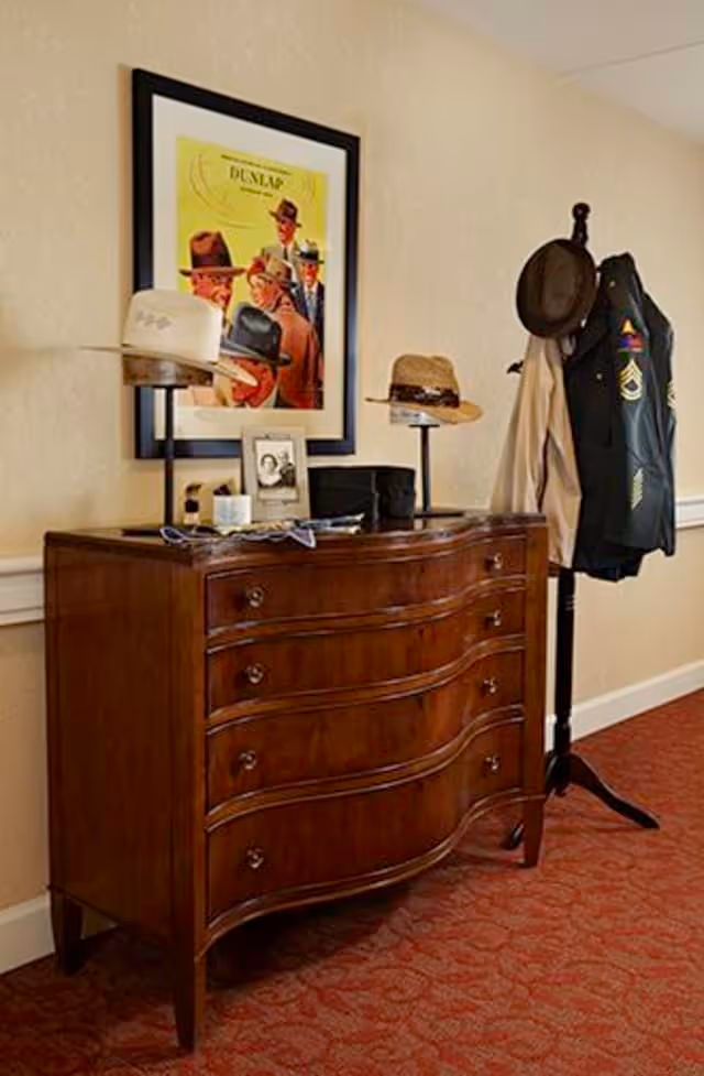 A wooden dresser with four curved drawers stands against a beige wall. On top of the dresser are several hats displayed on stands, a framed black and white photo, and some small items. Above the dresser hangs a framed vintage-style poster featuring men wearing hats. To the right of the dresser is a coat rack holding a hat, a beige coat, and a dark jacket with military-style patches. The floor is covered with a red patterned carpet.