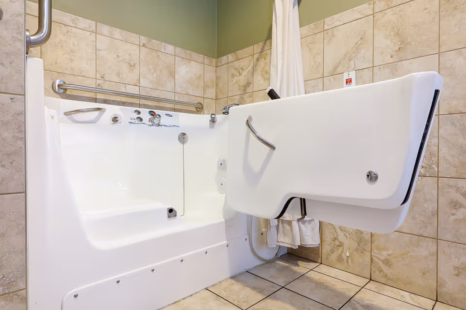 A walk-in bathtub with a side door open, featuring grab bars and control knobs, surrounded by beige tiled walls and floor.
