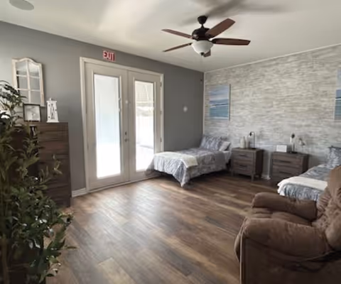 A bright and spacious bedroom with two single beds, each with gray bedding and pillows. Between the beds are two matching nightstands with lamps and a small decorative item. The room has wooden flooring, a ceiling fan with light, and a large glass door with an exit sign above it. There is a brown recliner chair in the foreground and a tall dresser with decorative items on the left side near a green plant. The walls are painted gray with one accent wall featuring a textured design and two framed pictures.
