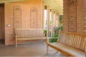 Covered outdoor patio area with two wooden benches placed against brick columns and walls, with decorative metal wall art and some greenery visible in the background.