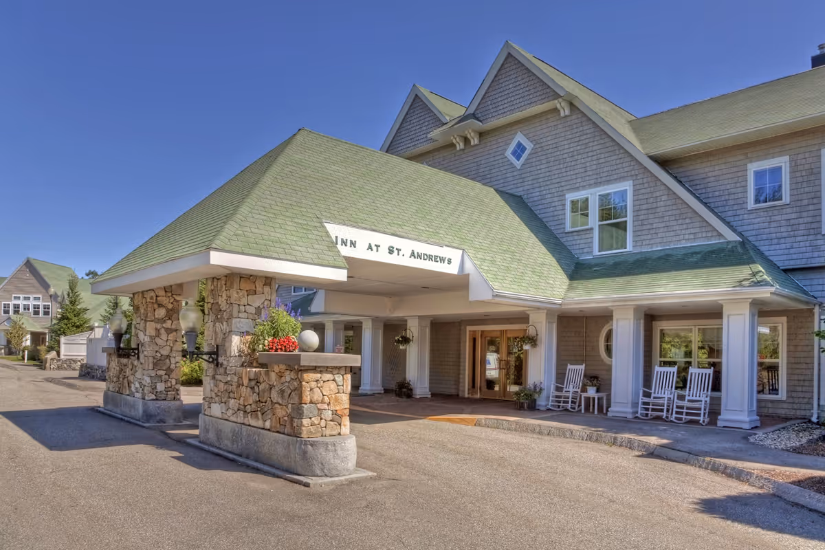 Exterior view of the Assisted Living at St. Andrews Village showing the entrance with a green roof, stone pillars, and white rocking chairs on the porch under a clear blue sky.