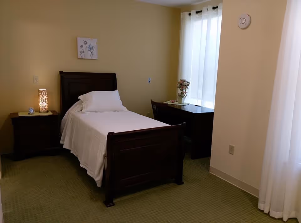 A small bedroom with a single dark wooden bed covered with white bedding and a pillow. Next to the bed is a matching nightstand with a decorative lamp and a small clock. A dark wooden desk with a chair is positioned near a window with sheer white curtains, and a small flower arrangement is on the desk. The walls are painted light yellow, and there is a small framed artwork above the bed.