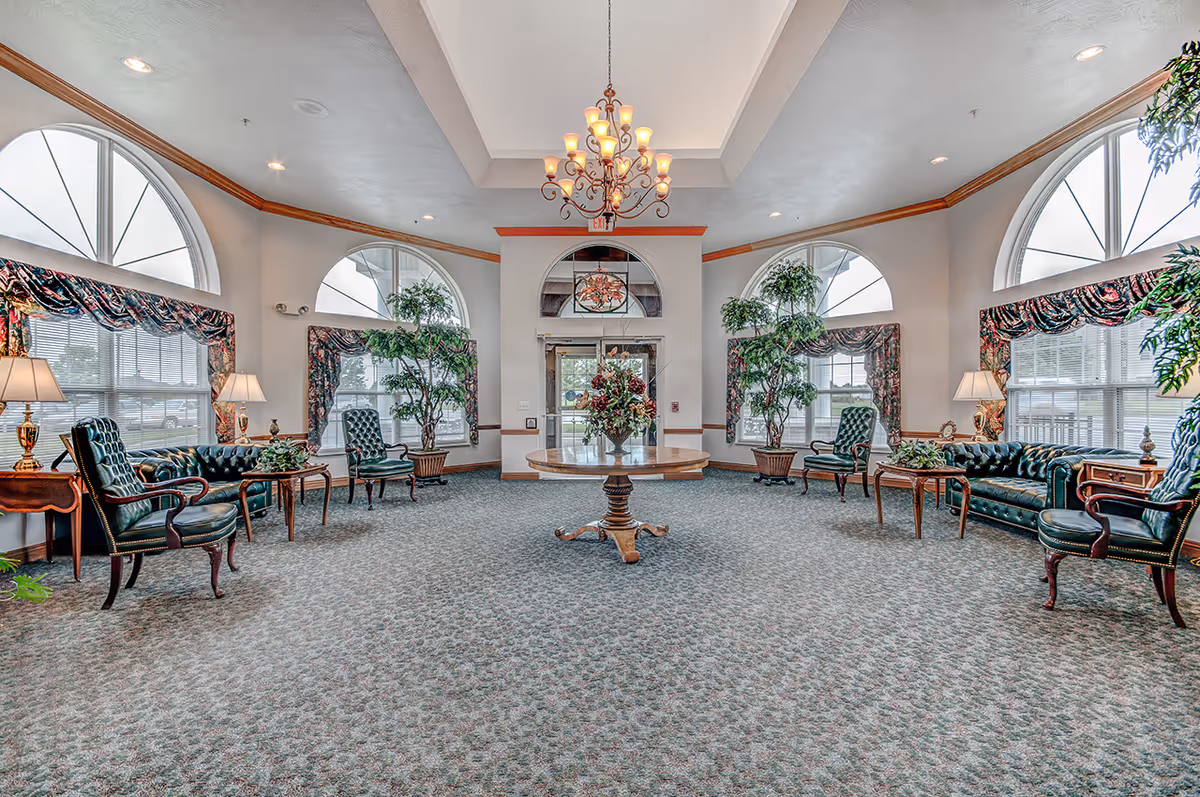 A spacious, well-lit living room with large arched windows, green leather armchairs and sofas arranged around wooden side tables, a central round wooden table with a floral arrangement, potted plants, and a chandelier hanging from the ceiling.