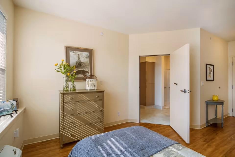 A bright bedroom with a wooden floor, a bed with a blue blanket in the foreground, a chest of drawers with a vase of yellow flowers and a framed picture of the Eiffel Tower on the wall above it. There is an open door leading to another room with tiled flooring, and a small side table with a yellow pitcher and a framed picture on the wall beside it.