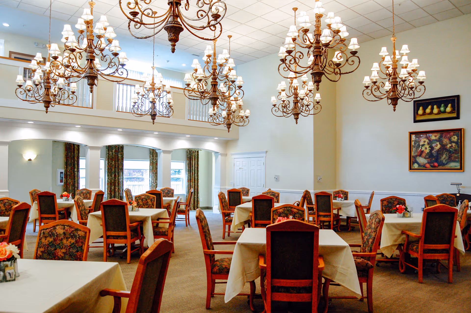 A spacious dining room with multiple tables covered in white tablecloths and surrounded by wooden chairs with floral upholstery. Several ornate chandeliers hang from the ceiling, and the room features large windows with curtains, light-colored walls, and framed artwork.