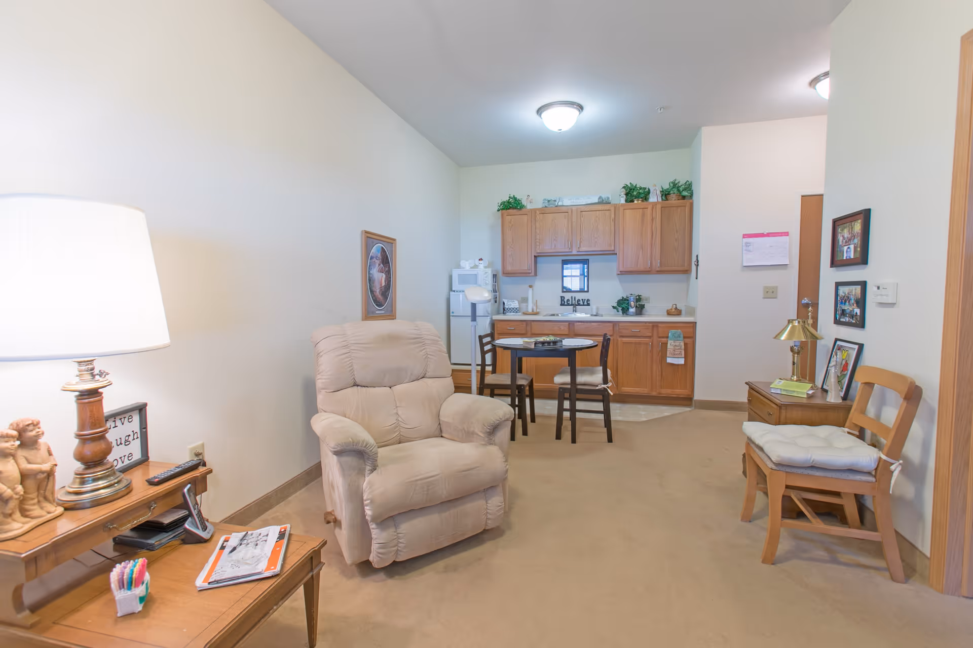 A cozy living area in a senior living facility featuring a beige recliner chair, a small wooden table with a lamp and decorative items, a wooden chair with a cushion, and a kitchenette with wooden cabinets, a small dining table with two chairs, and various kitchen appliances and decor.