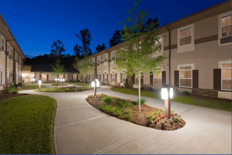 Night view of a senior living facility courtyard with well-lit pathways, small trees, landscaped garden beds, and a two-story building surrounding the courtyard.