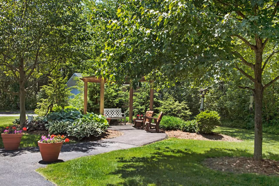 A peaceful outdoor garden area with a paved pathway, two large potted plants with flowers, a wooden pergola with a white bench underneath, and two wooden chairs. The area is surrounded by green trees, bushes, and well-maintained grass.