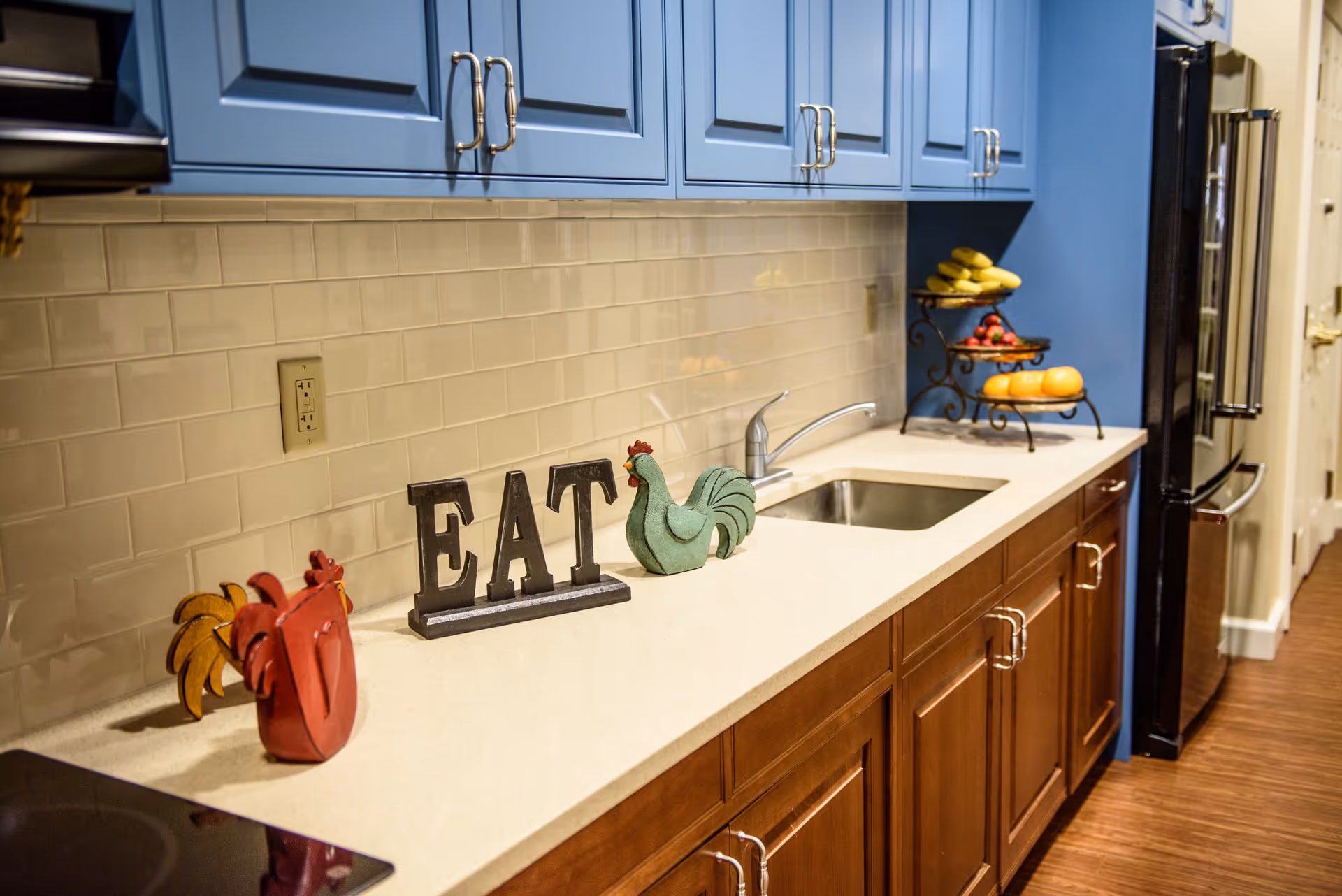 A kitchen countertop with blue upper cabinets and wooden lower cabinets. On the counter, there are decorative items including a red rooster, a green rooster, and black letters spelling 'EAT'. There is a sink with a faucet, a tiered fruit stand holding bananas, strawberries, and oranges, and a black refrigerator at the end of the counter.