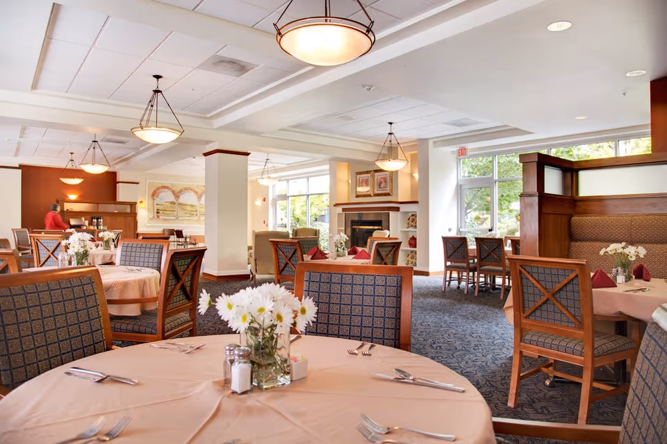 Bright dining room with round tables set with tablecloths and flower centerpieces, wooden chairs, and large windows.