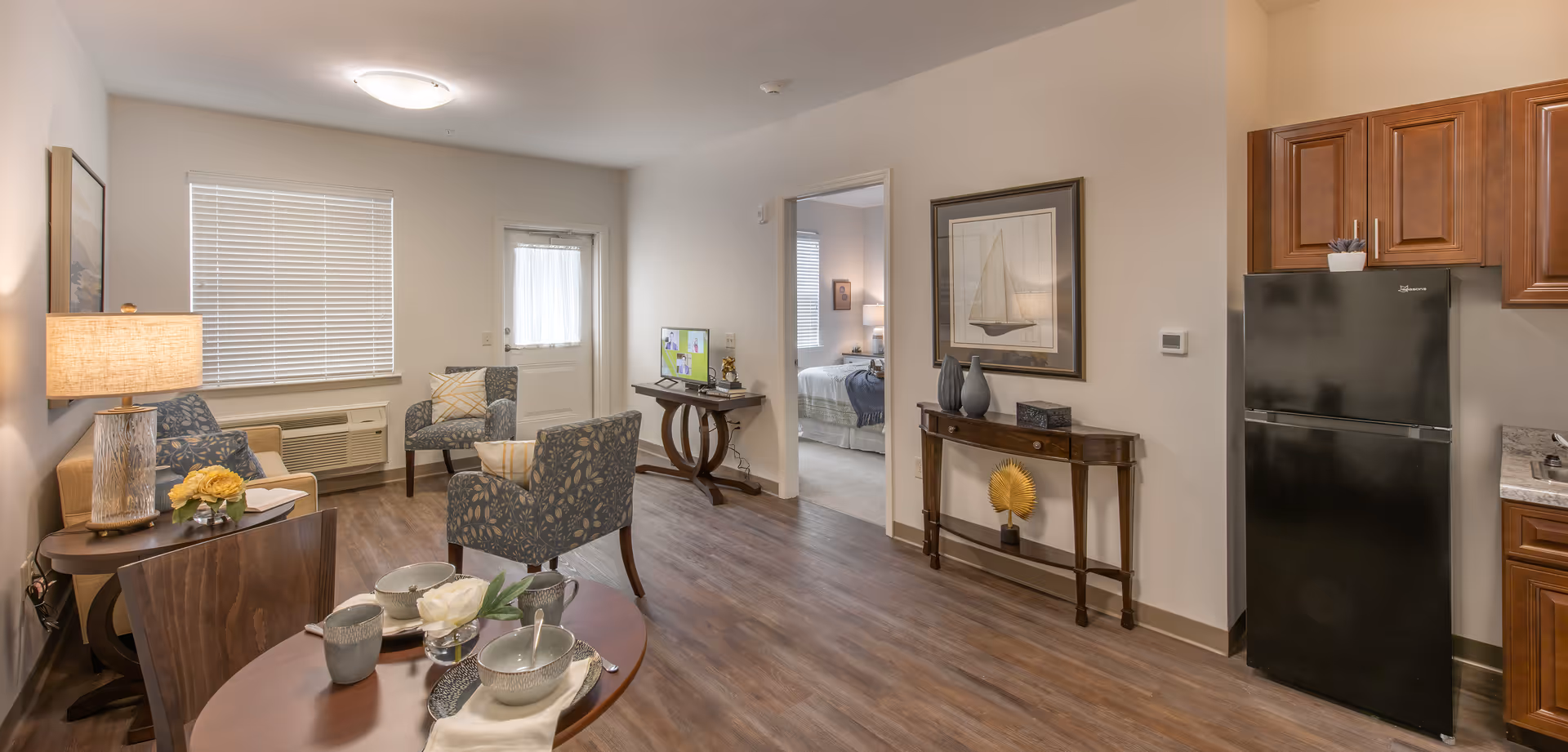 Bright furnished living room with chairs and a small dining table, a kitchenette with a black refrigerator, and a doorway leading to a bedroom.